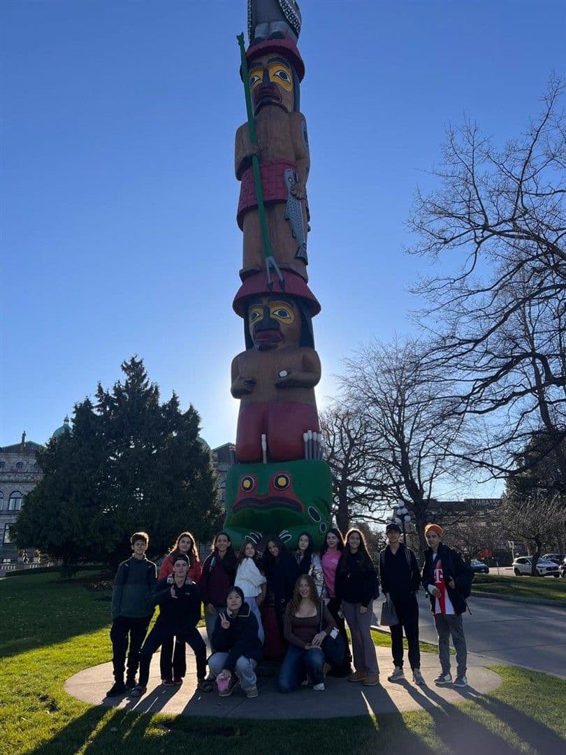 Alunos reunidos ao pé de um totem indígena entalhado, em dia de céu azul
