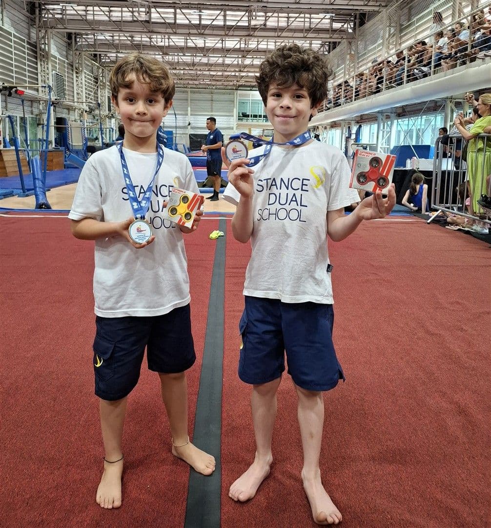 Dois alunos com camiseta Stance Dual School mostram medalhas e prêmios no ginásio de ginástica