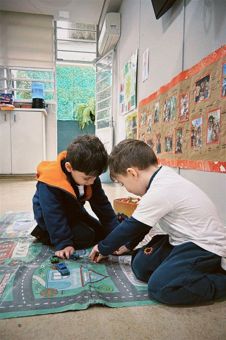 Dois meninos ajoelhados num tapete de pista, brincando juntos com carrinhos na sala