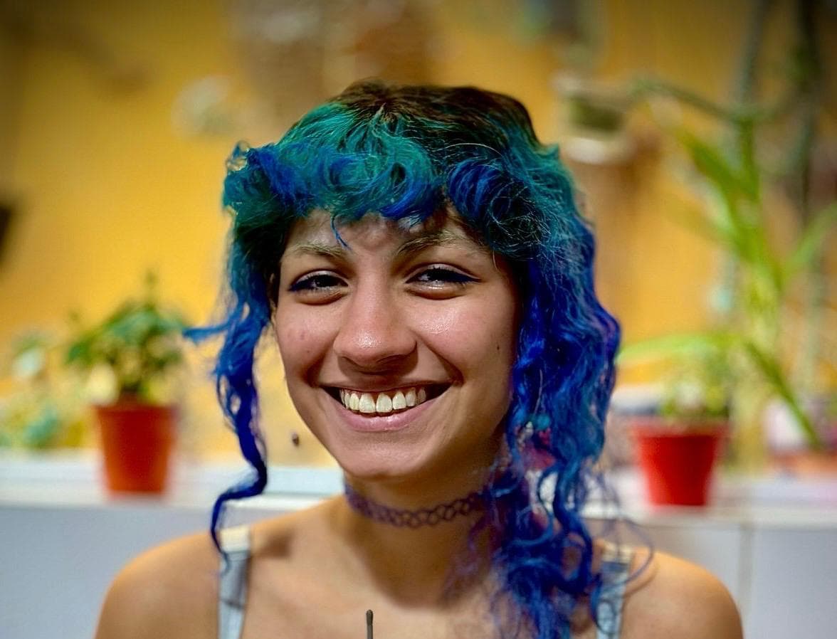 Retrato de Mayara Reyes, sorridente, de cabelos cacheados tingidos de azul e verde