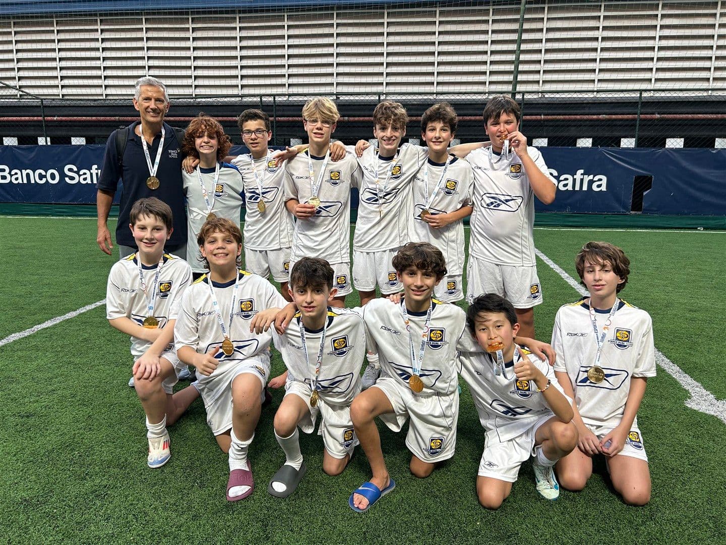 Time de futebol da Stance, de uniforme branco, posa com medalhas no campo ao lado do técnico
